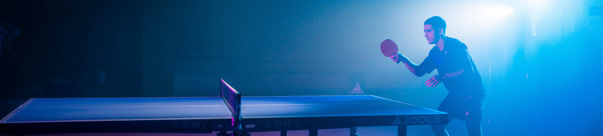 Man playing table tennis with a racket at a JOOLA table in dramatic blue lighting
