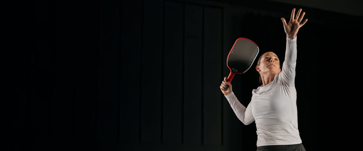 Woman in white athletic wear holding a red pickleball paddle against a dark background